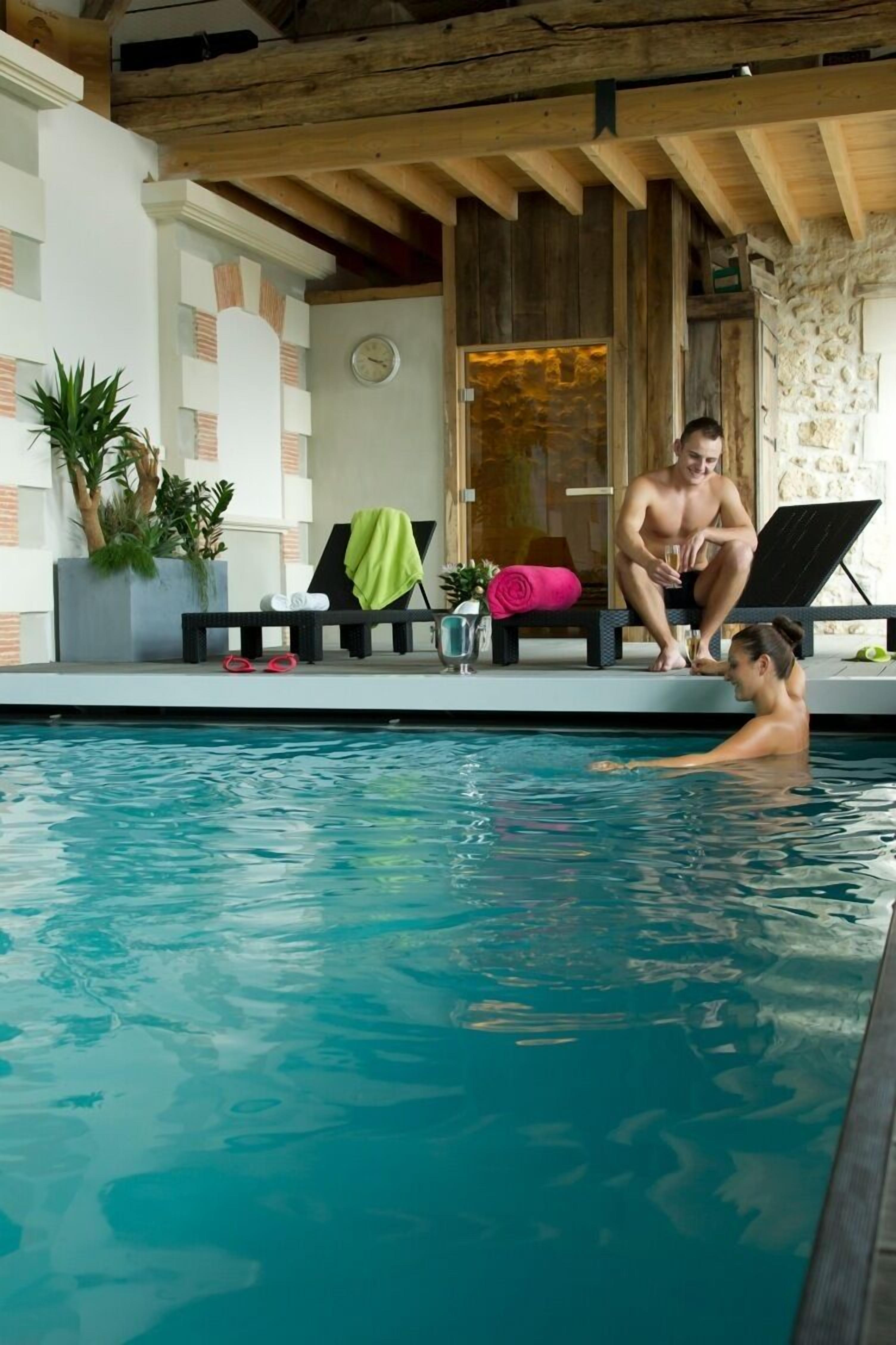 Une piscine intérieure avec un plafond en bois et des murs en pierre. On y trouve des chaises longues et un banc avec des serviettes.