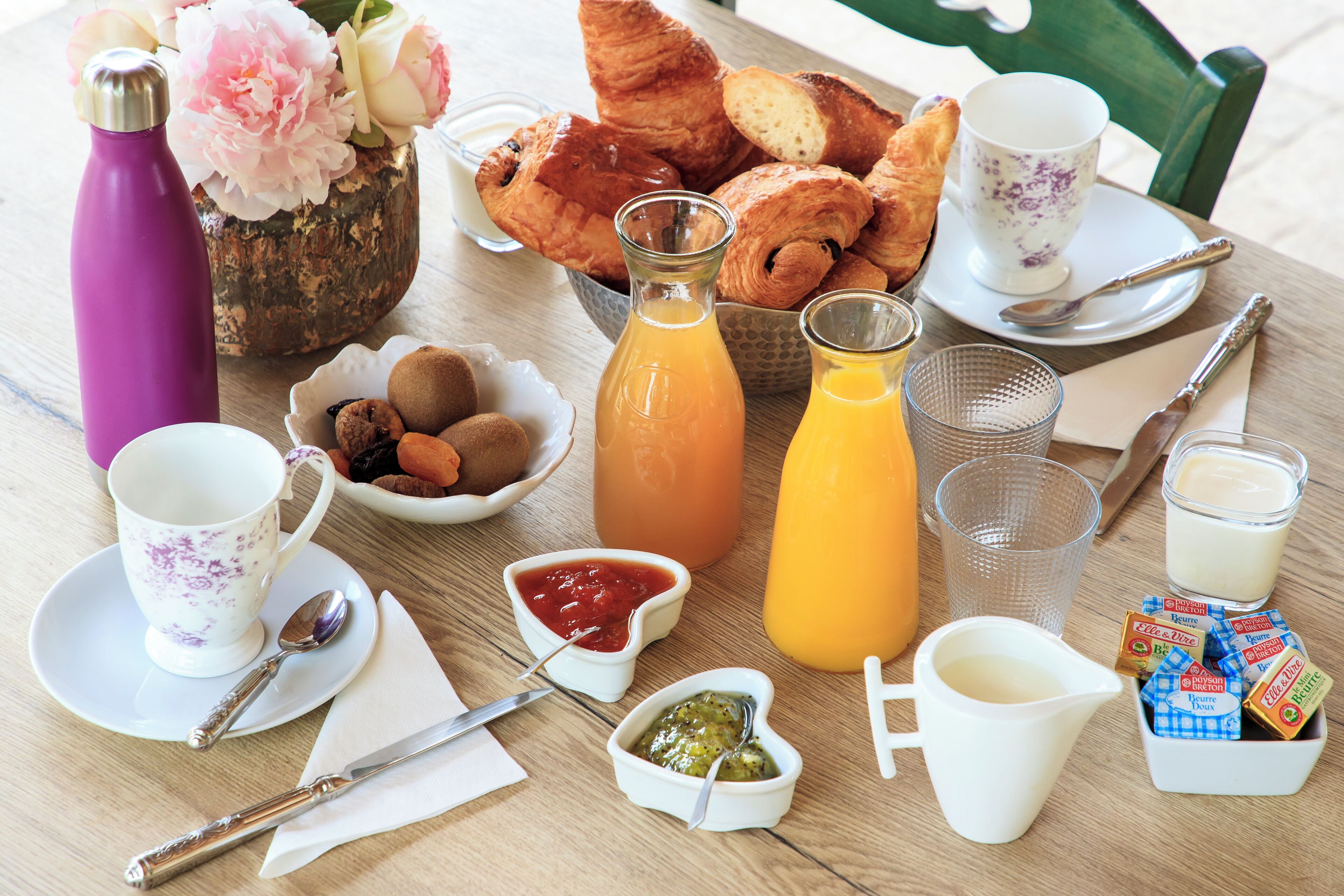 Ein Frühstückstisch mit Croissants, Gebäck, einem Thermosbehälter, einem Krug Orangensaft, einer Tasse Kaffee, einer Schale Obst, Gewürzen und einer Schachtel Müsli.
