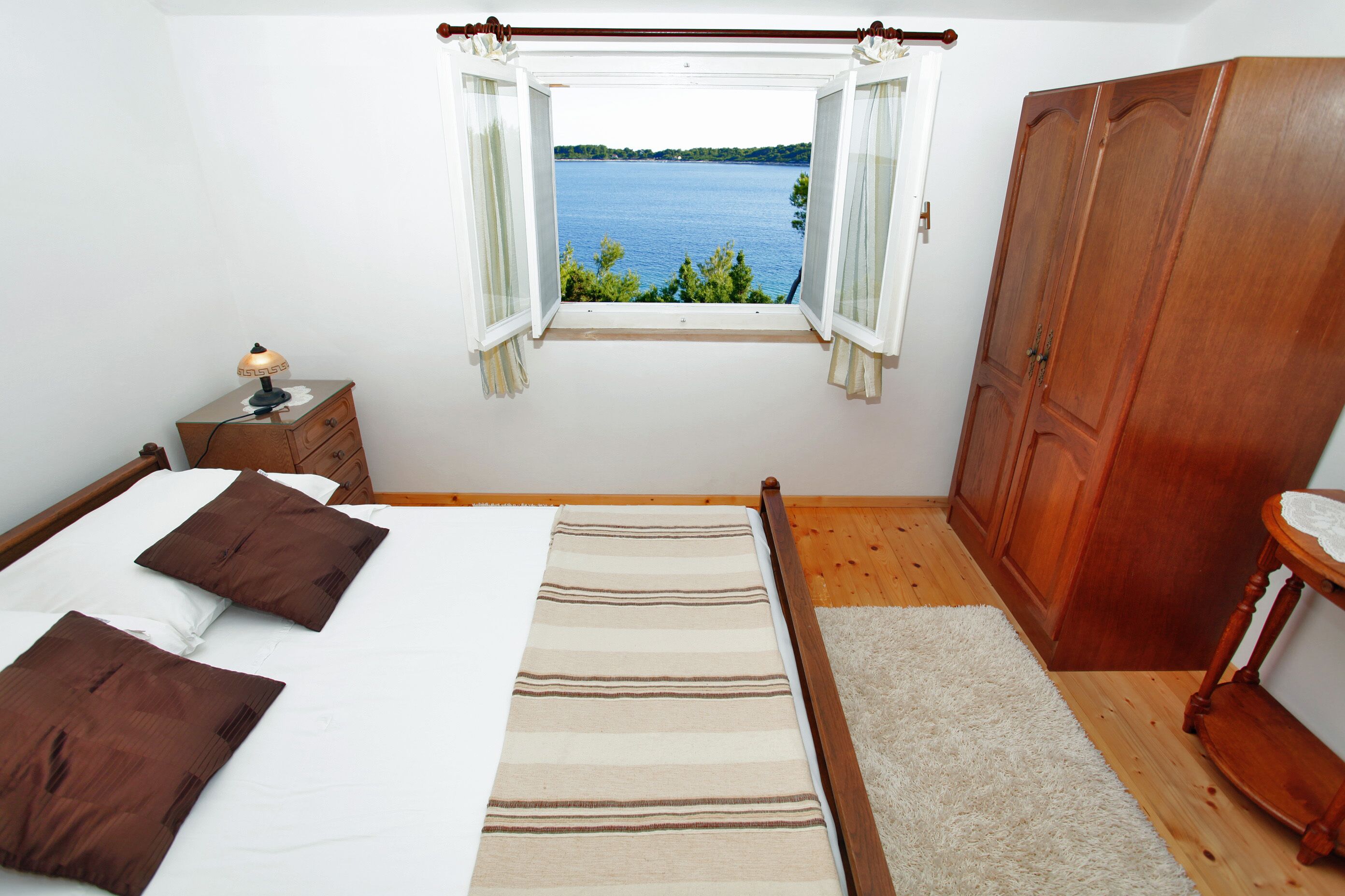 A bedroom with a bed, a nightstand, a wooden wardrobe, and a window with a view of a lake.