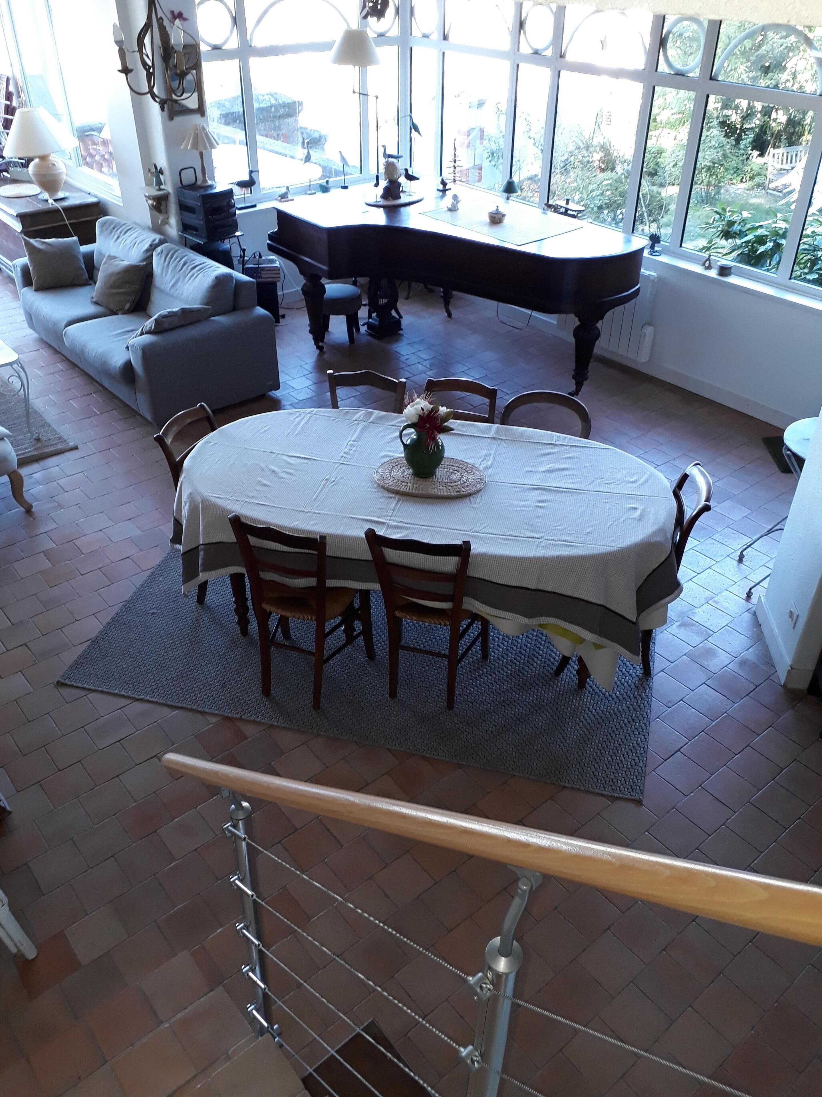 A dining area with a round table, chairs, and a vase of flowers. There is a sofa and a piano in the background.