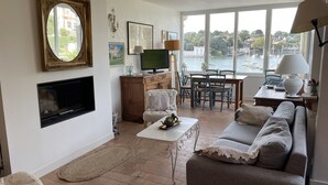 Ett vardagsrum med TV, öppen spis, matplats och stora fönster med utsikt över en sjö eller ett annat vattendrag.