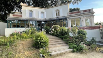 Un bâtiment à deux étages doté d’une entrée couverte, d’un jardin agrémenté de fleurs roses et d’un escalier en pierre menant à l’entrée.