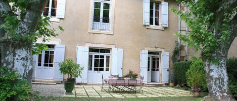 A two-story building with white shutters, a garden with a table and chairs, and potted plants.