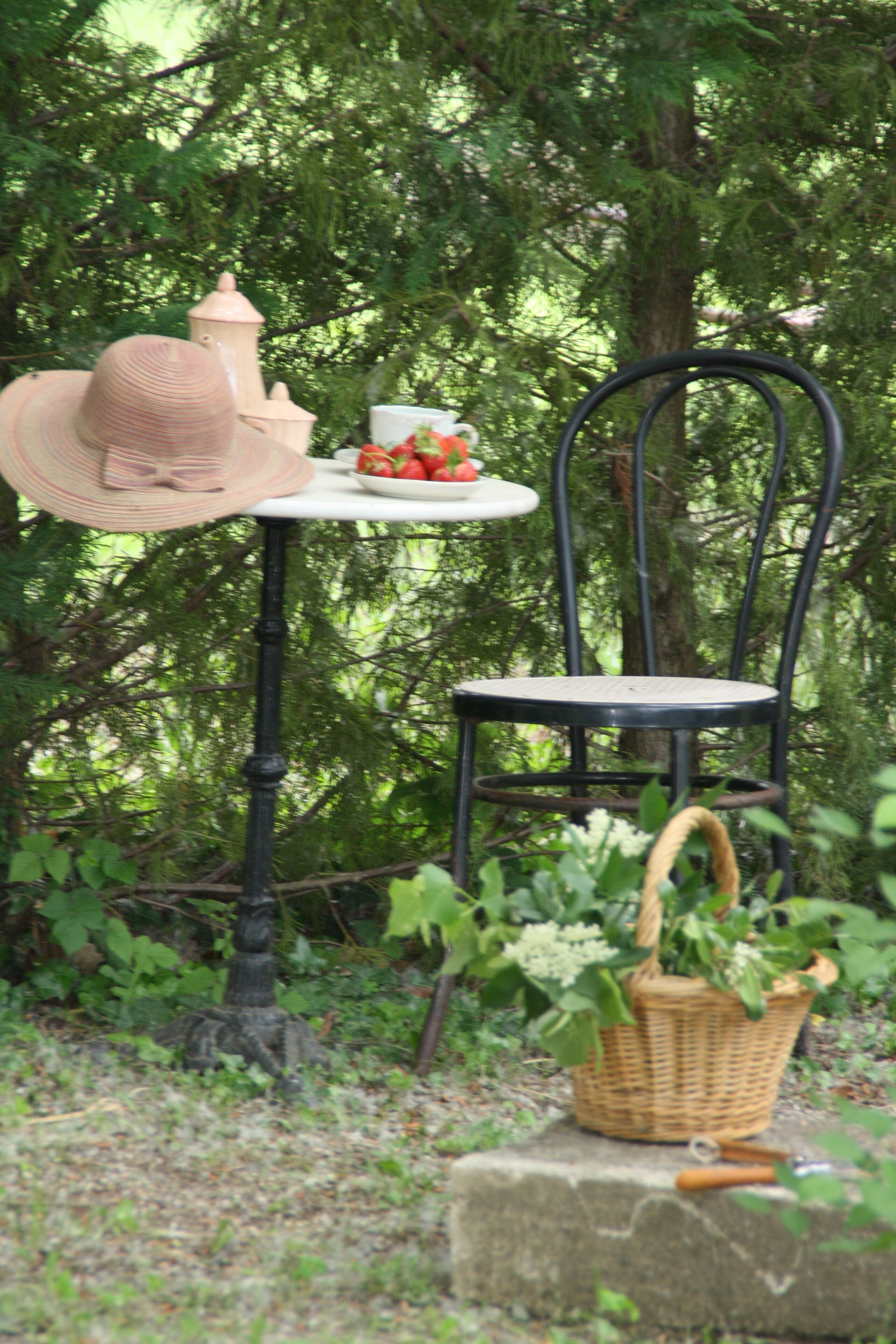 A rustic outdoor setting with a wicker basket on a stone, a black metal chair, a white table with a hat and a bowl of fruit, and a lamp post in the background.