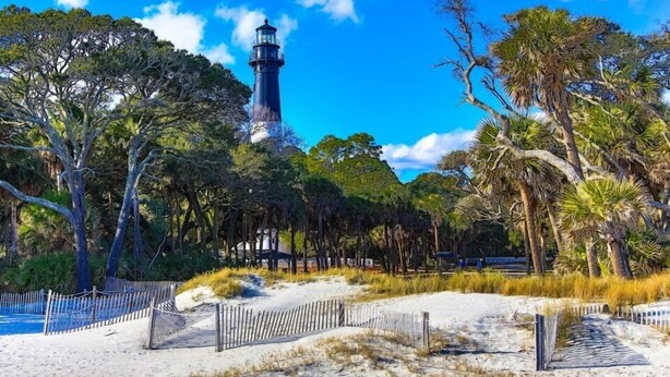Un phare entouré d’arbres et une zone sablonne délimitée par une clôture.