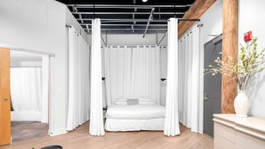 Un dormitorio con cama, cortinas blancas, suelo de madera, una mesita y un aparador.