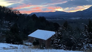A snow-covered house with a deck, surrounded by trees and mountains in the background.