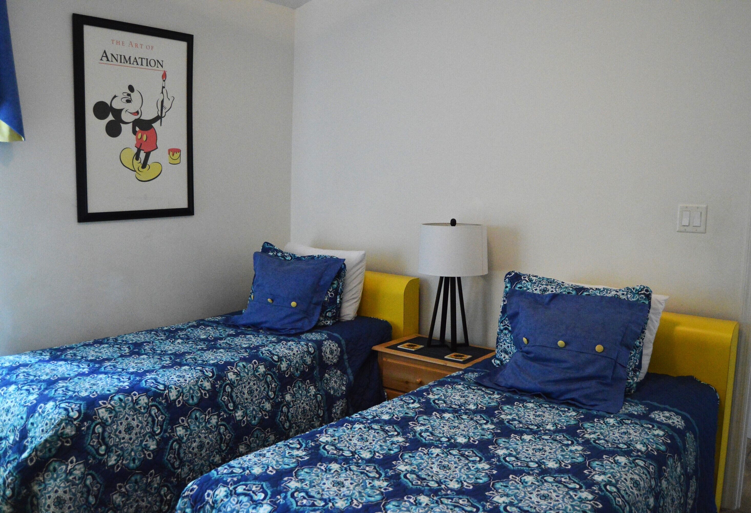 Une chambre avec deux lits, chacun recouvert d’une courtepointe bleue à motifs et agrémenté d’oreillers assortis. Un tableau encadré représentant Mickey Mouse est accroché au mur.