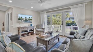 A living room with a ceiling fan, a large window with curtains, a sofa, a coffee table, and a TV mounted on the wall. - Award-Winning Resort Amenities | Racquet Club Villa 6 (Isle of Palms)