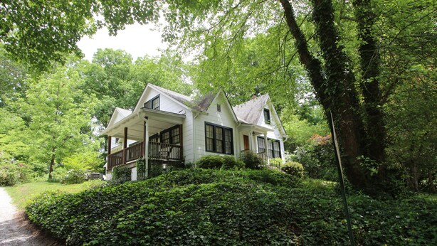 Une maison blanche dotée d’un porche, entourée d’une végétation luxuriante et d’arbres.