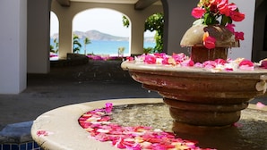 Una fuente con flores y pétalos rosados en un patio con arcos y vista al mar.