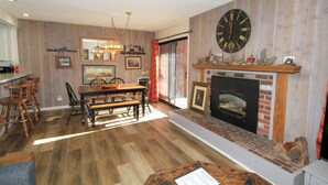 A dining area with a wooden table and chairs, a fireplace with a clock above it, and a kitchen in the background. - Mammoth Ski & Racquet Club #114 -Pet Friendly (Mammoth Lakes)