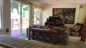 Ein gemütliches Wohnzimmer mit einer Couch, einem Couchtisch und einem Fernseher. Ein großes Fenster bietet einen Blick ins Freie.