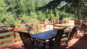 Eine Terrasse im Freien mit Tisch und Stühlen, umgeben von einem Holgitter und Blick auf ein bewaldetes Gebiet.