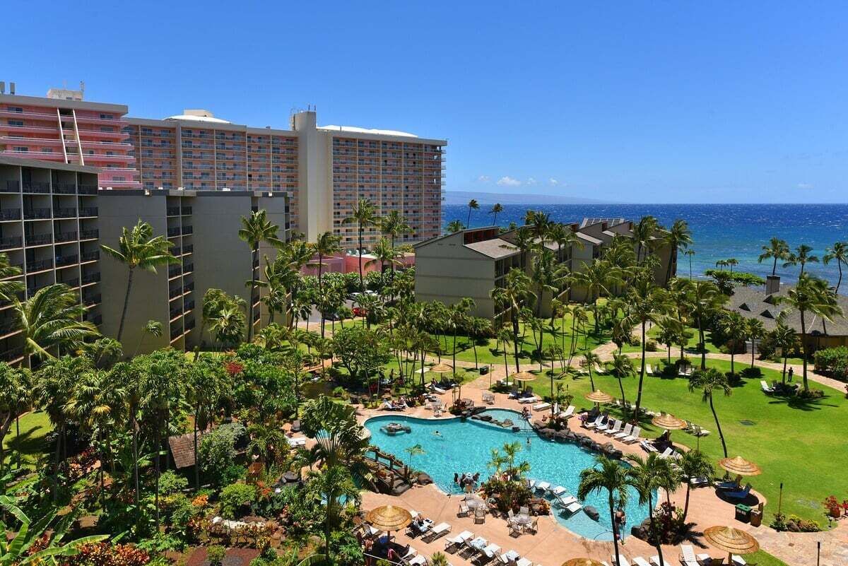 A resort with a large pool surrounded by palm trees and lounge chairs, overlooking a beach.