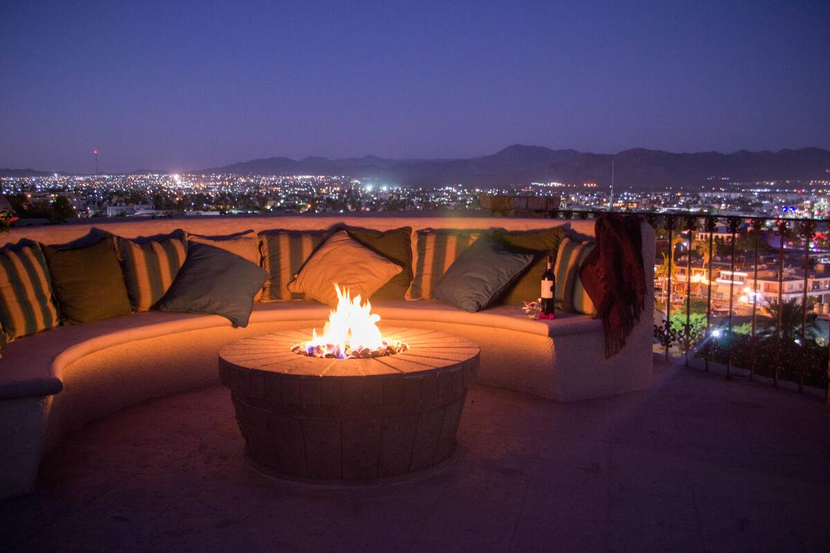 A rooftop seating area with a circular fire pit, cushioned seating, and a cityscape view in the background.