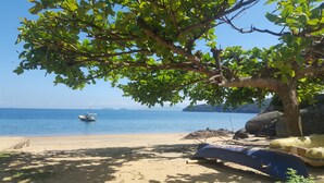 A beach with a boat on the water, a tree providing shade, and a sandy area with some objects on the ground. - Pousada Três Coqueiros (Angra dos Reis)