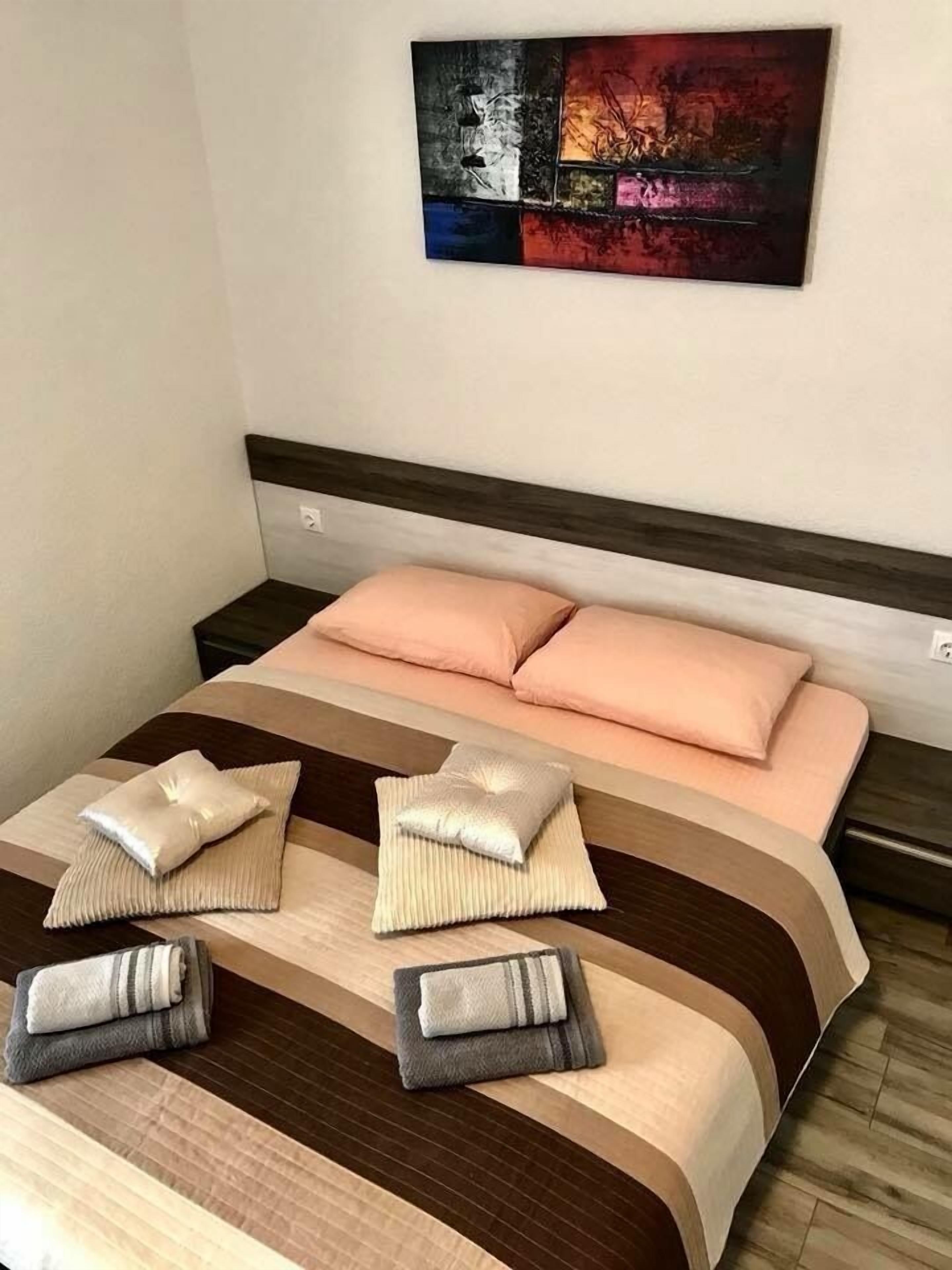 A neatly made bed with a striped bedspread and two pillows, a small bedside table with a lamp, and a colorful abstract painting on the wall.