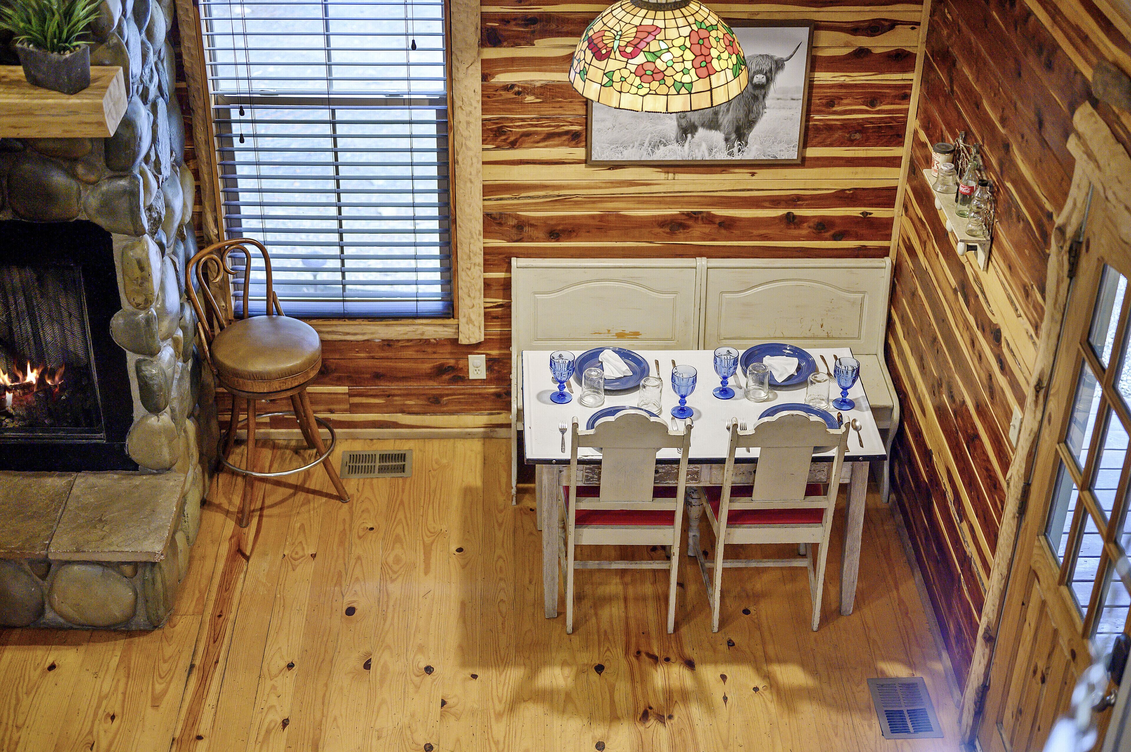 Interior rústico de madera con chimenea de piedra, una mesa de comedor para dos personas y un ventanal con persianas.