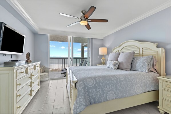 A bedroom with a bed, a ceiling fan, a TV, a dresser, and a balcony with a view.