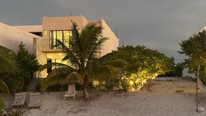 A modern beach house with a balcony, palm trees, and a sandy beach.