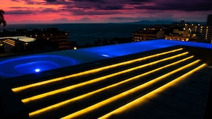 Une piscine sur le toit, avec des marches illuminées, offrant une vue sur l’océan au coucher du soleil.