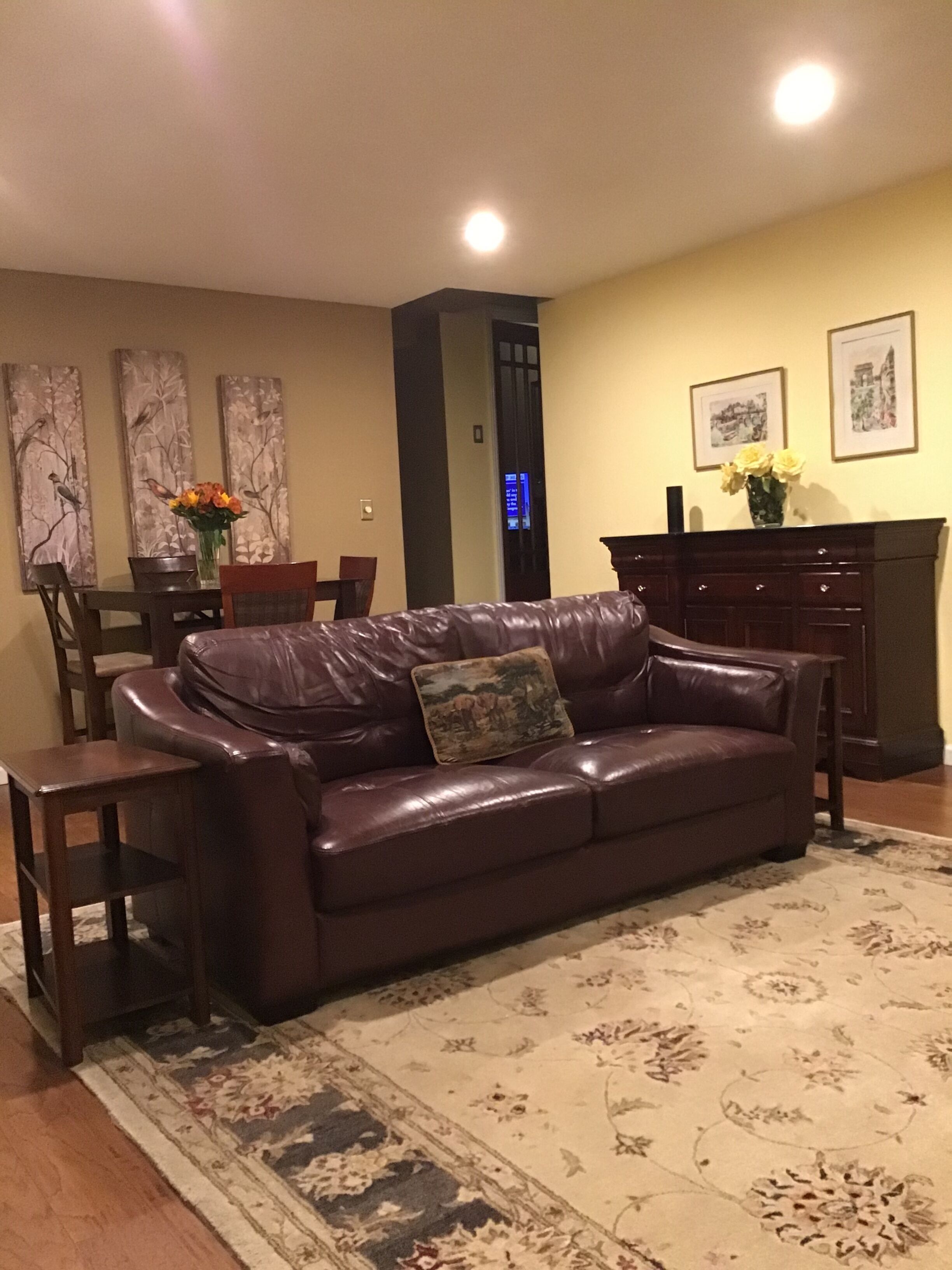 A living room with a brown leather sofa, a dining table with chairs, a vase with flowers, and artwork on the walls.