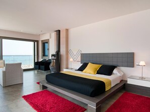 A modern hotel room with a large bed, a seating area with chairs, a view of the sea, and a fireplace. - Bohemi, luxury seafront villa (Santa Margalida - Can Picafort/Son Serra Marina)