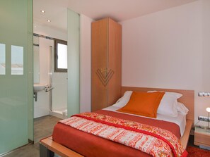 Habitació d'hotel amb un llit, una taula de nit, un armari de fusta, una porta de vidre i un bany visible a través d'una porta oberta.