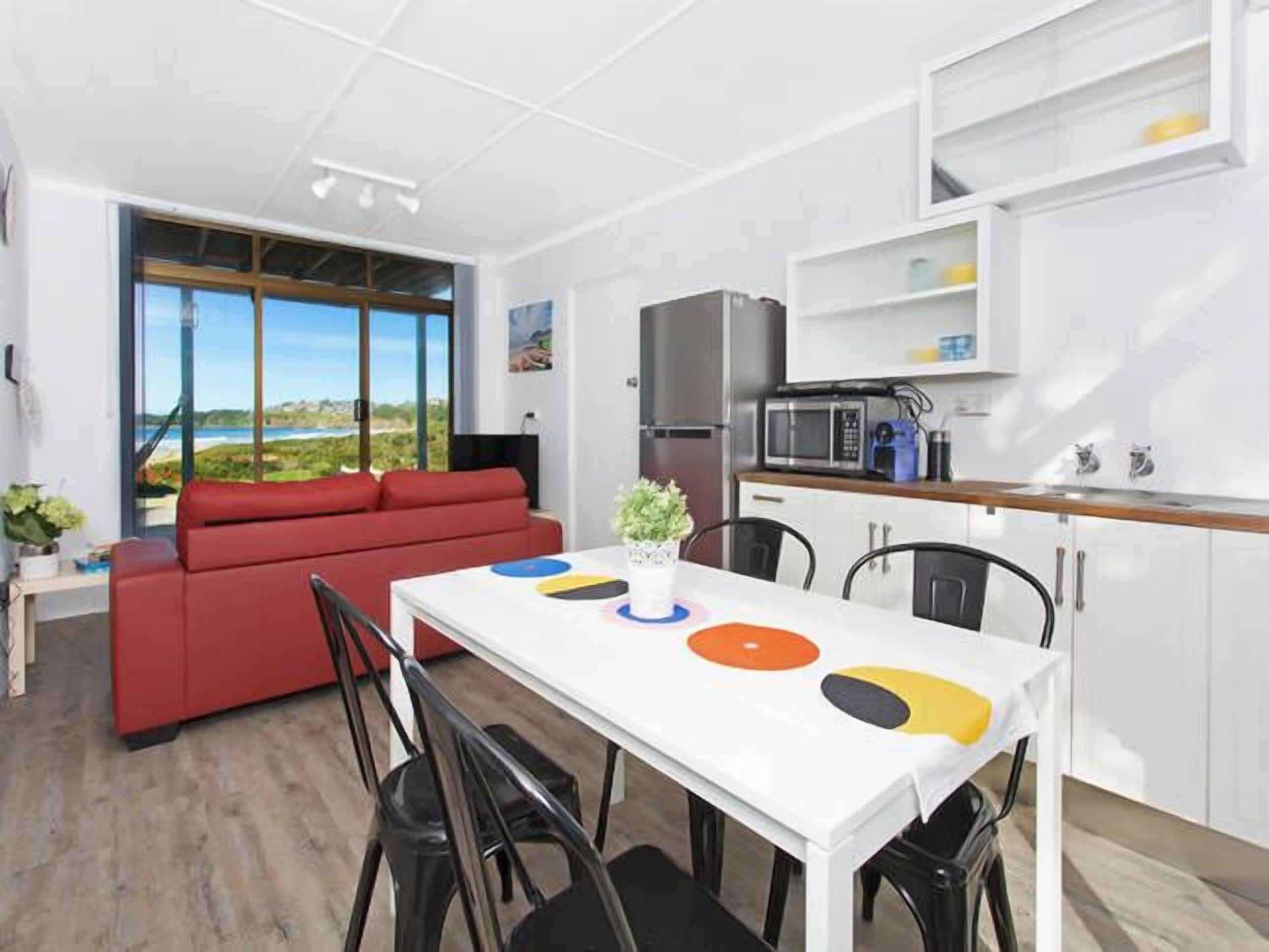 A modern living room with a red sofa, a dining table with a white tablecloth, and a kitchen area with a microwave and cabinets.