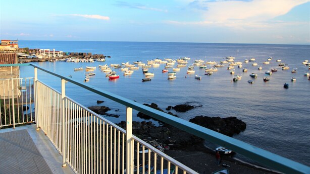 A coastal view with a marina filled with boats, a rocky shoreline, and a building with a red roof in the background.