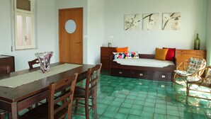 A room with a wooden dining table, chairs, a sofa with colorful cushions, a bed, and a green-tiled floor.