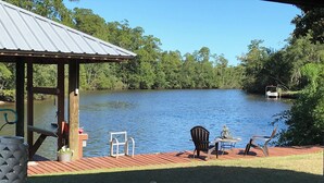 Una terraza de madera con techo de metal que da a un lago, una mesa con sillas y un bote al fondo.