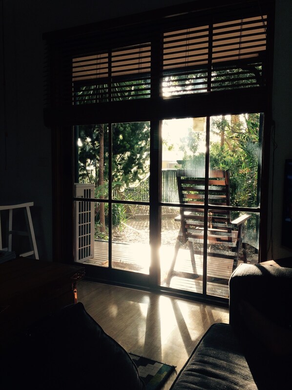 A room with a large window, wooden blinds, a wooden deck, a chair, and a table. - Spacious and unique beach cottage for relaxing getaways (Barwon Heads)