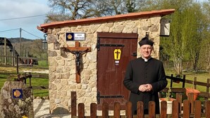 A stone structure with a wooden door, a cross, and a sign with a shell logo. - Stone Cottage The Compostelana (Curtis)