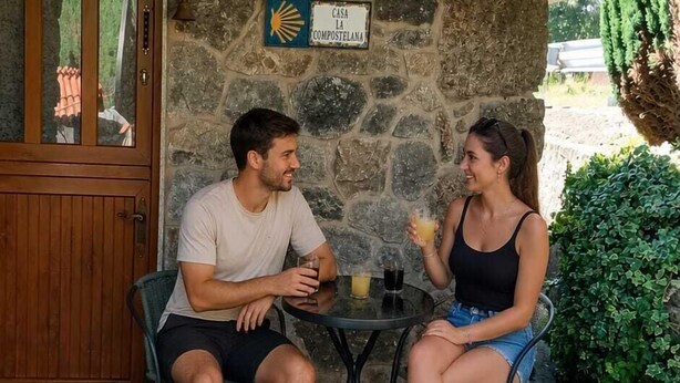Two people seated at a table under a wooden roof, with a stone wall and a door with a glass panel in the background. - Stone Cottage The Compostelana (Curtis)