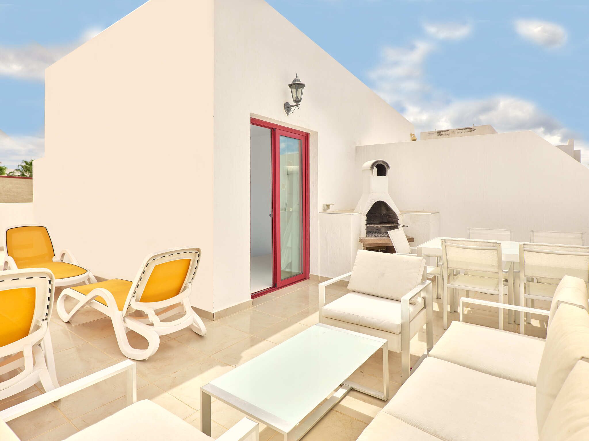 Une terrasse sur le toit, dotée de mobilier blanc, d’un four à pizza et d’une porte vitrée donnant accès à un espace intérieur.