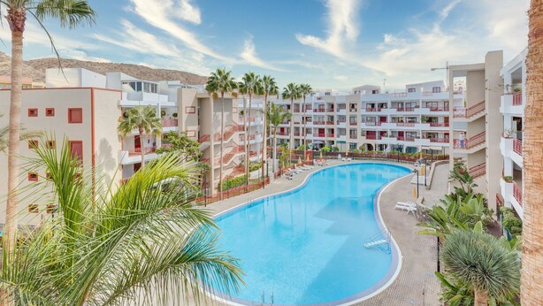 A resort with a large swimming pool surrounded by palm trees and a multi-story building with balconies.