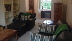 Een woonkamer met een leren bank, een gestreept fauteuil, een houten salontafel, een boekenkast, een houten kast en een raam met gordijnen.