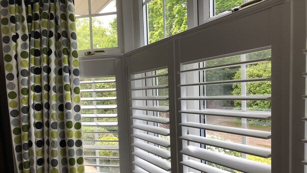 Een kamer met een erker, witte luiken, uitzicht op groen en een gordijn met stippen.