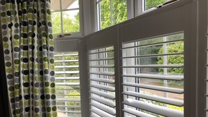 A room with a bay window featuring white shutters, a view of greenery outside, and a curtain with a polka dot pattern.