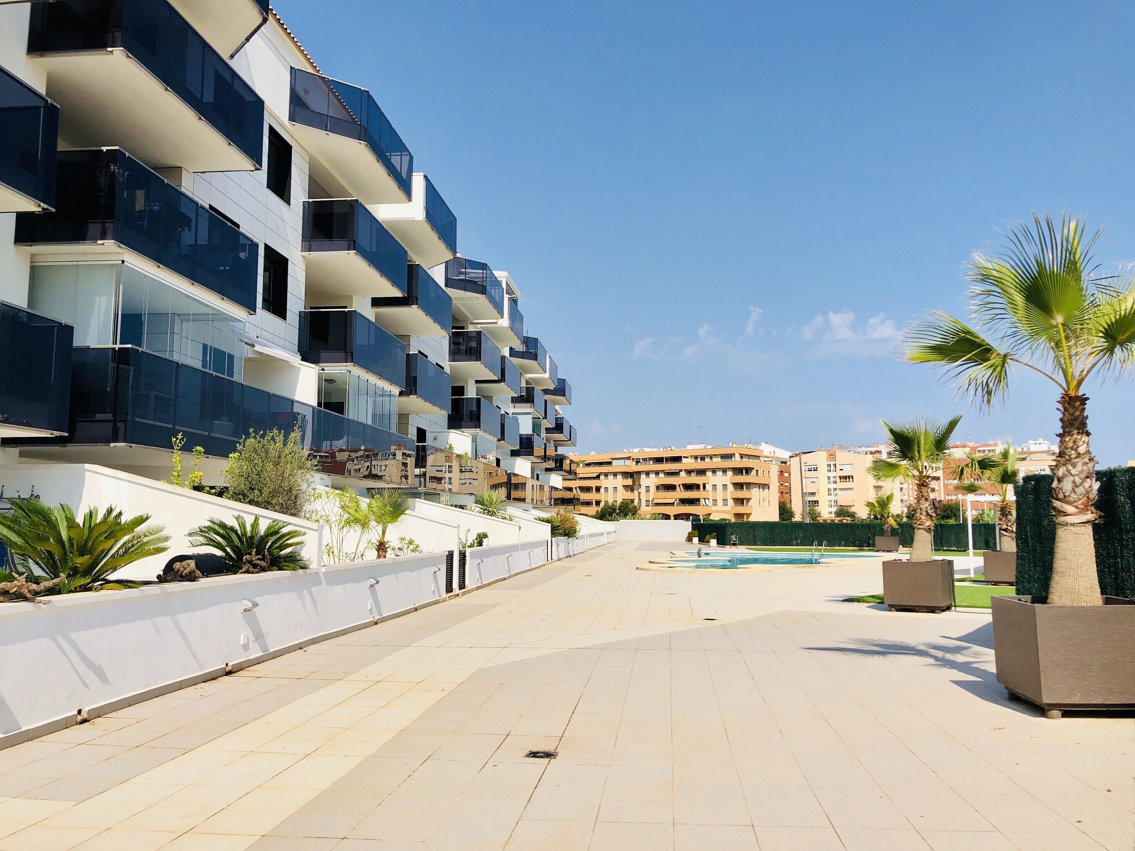 Un edificio residencial moderno con varios balcones, un camino pavimentado y palmeras.