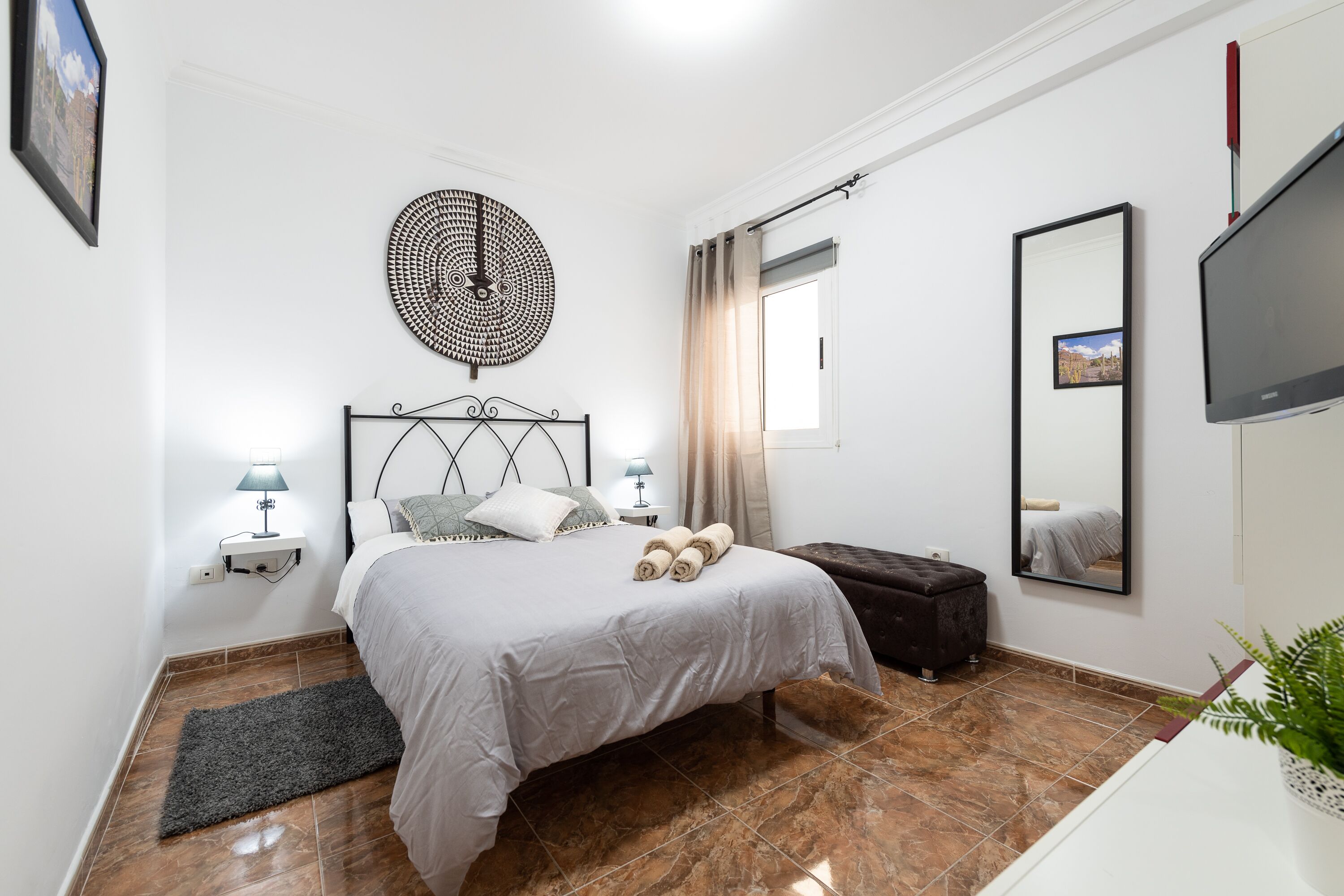 A bedroom with a bed, bedside lamps, a round wall decoration, a window with curtains, a flat-screen TV, and a mirror.