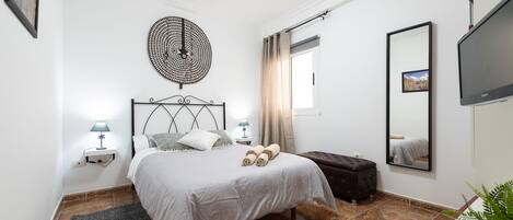 A bedroom with a bed, bedside lamps, a round wall decoration, a window with curtains, a flat-screen TV, and a mirror.