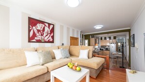 Ein modernes Wohnzimmer mit einem beigefarbenen Ecksofa, einem weißen Couchtisch mit Früchten und einer Küche im Hintergrund.