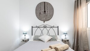 Ein ordentlich bezogenes Bett mit zwei Kissen, einem wandmontierten runden Dekorationsobjekt, zwei Nachttischlampen und einem Paar zusammengerollter Handtücher.