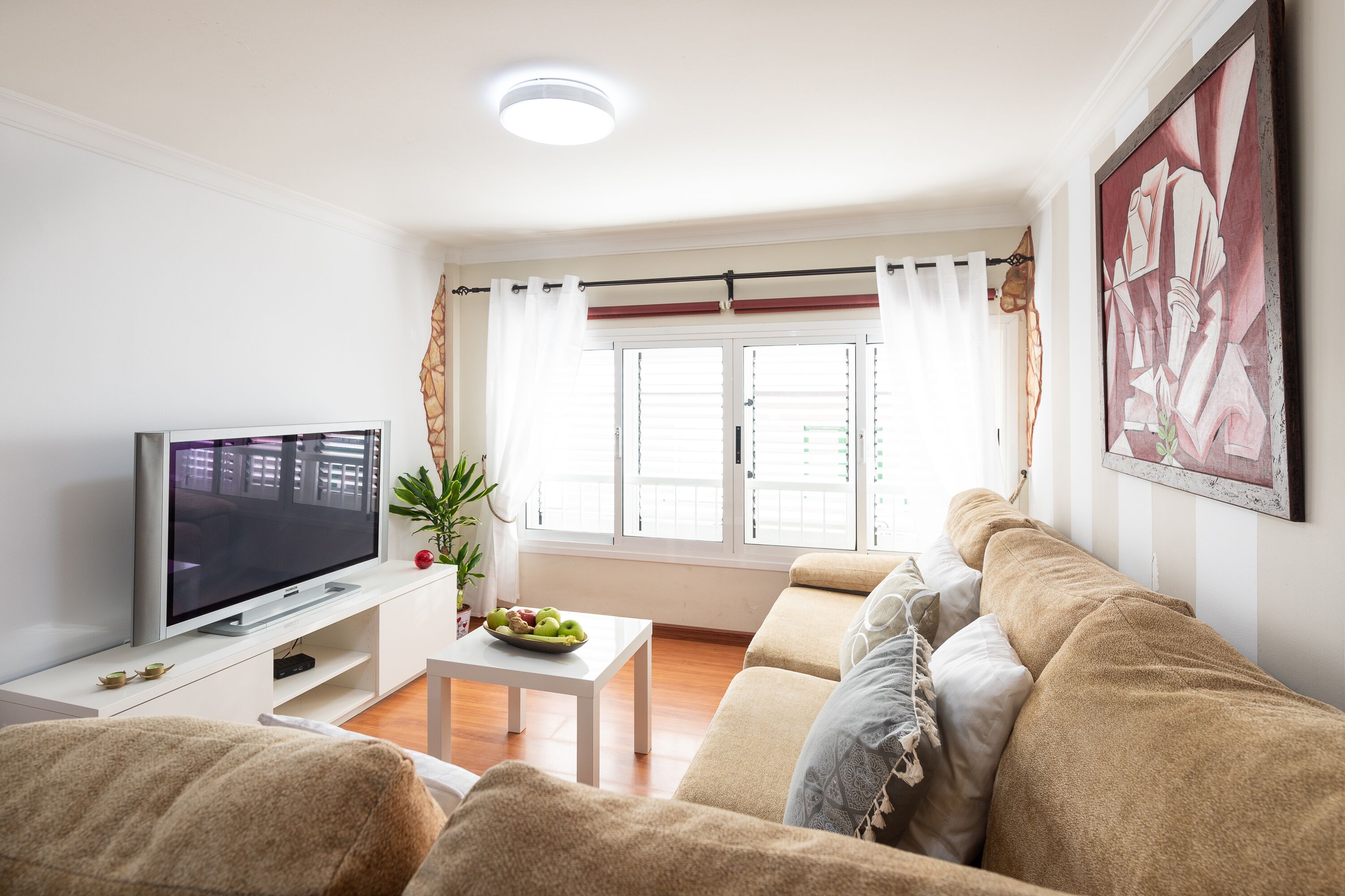 Una sala de estar con televisor de pantalla plana, una mesa de centro blanca, un sofá color beige y una ventana con cortinas.