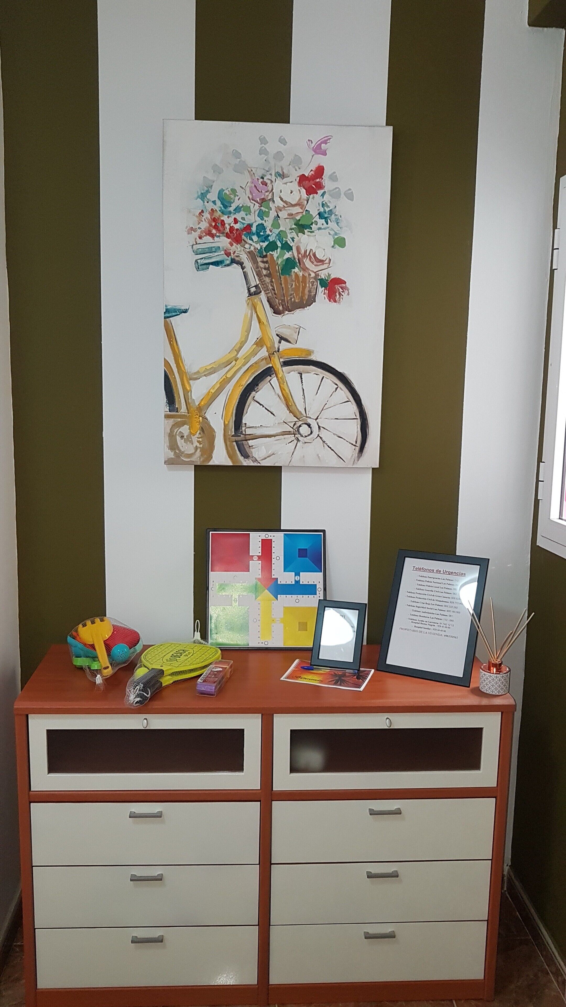 A room with a wall-mounted painting of a bicycle with flowers, a desk with drawers, a framed certificate, and a small table with various items.