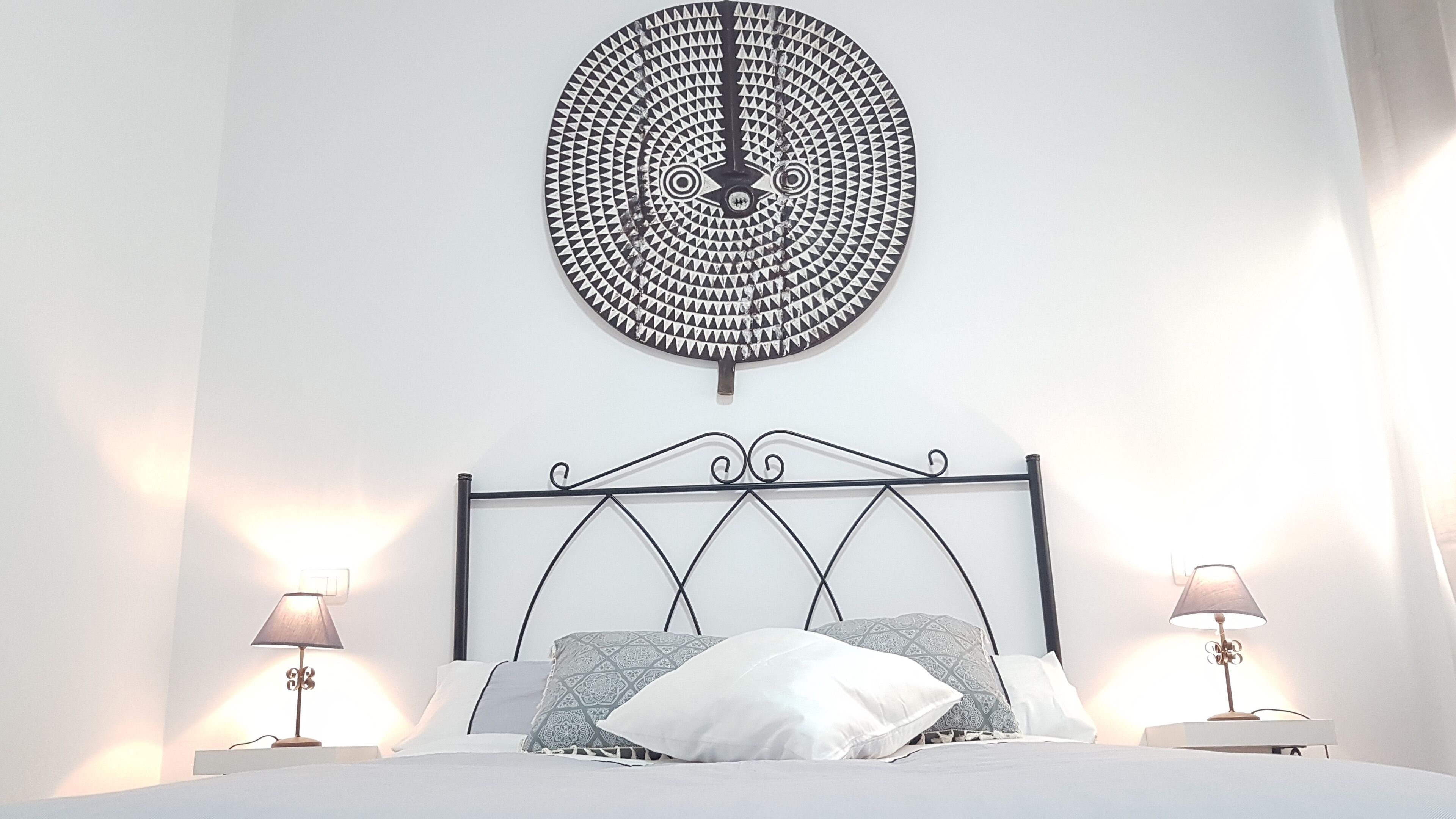 A bedroom with a bed, two bedside lamps, a decorative wall piece, and a metal headboard.