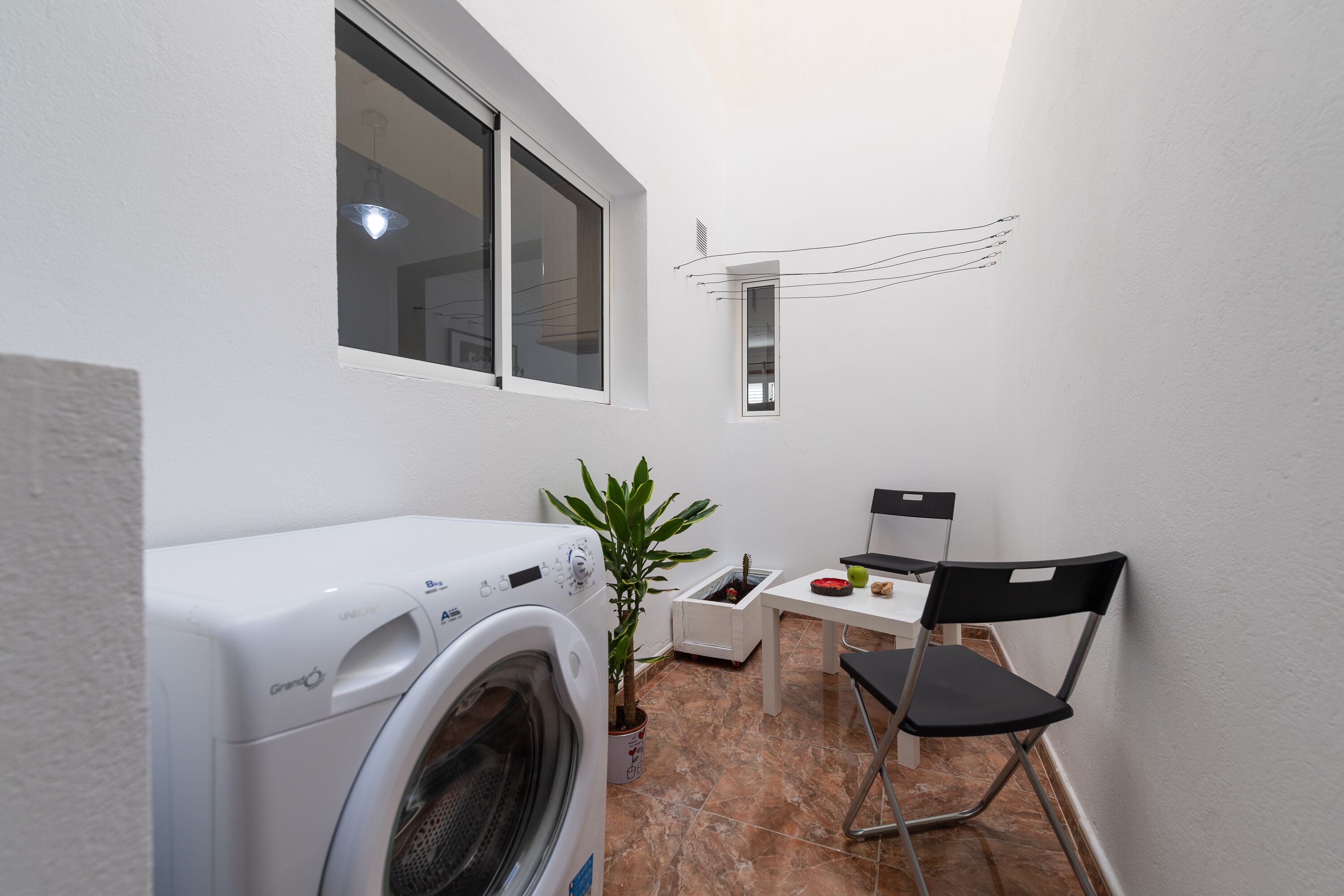 A compact laundry area with a washing machine, a small table, a chair, and a potted plant.